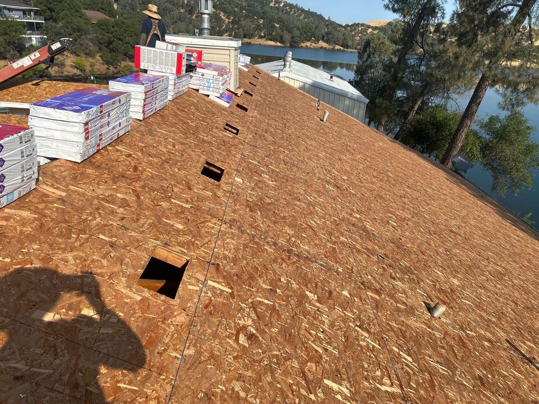 Full roof deck exposed with new OSB sheathing and GAF shingle bundles staged, lake visible